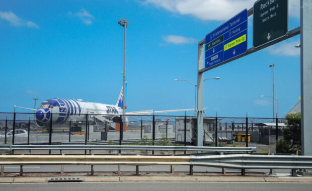 Airplane with R2-D2 livery is parked at Sydney Airport near a highway. The sky is clear, and nearby road signs point to the International Terminal.