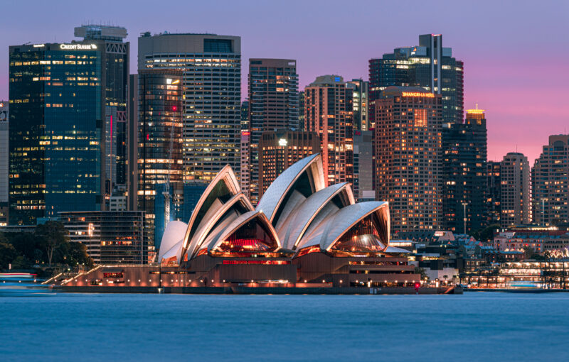 Sydney Opera House at dusk, illuminated against a vibrant city skyline. The sky is a gradient of pink and purple, reflecting off the calm harbor waters.