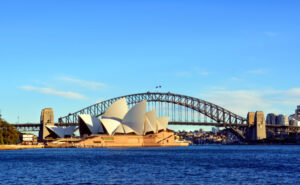 Sydney Opera House with its distinctive sails is set against the backdrop of the Sydney Harbour Bridge. The sky is clear and the water sparkles.