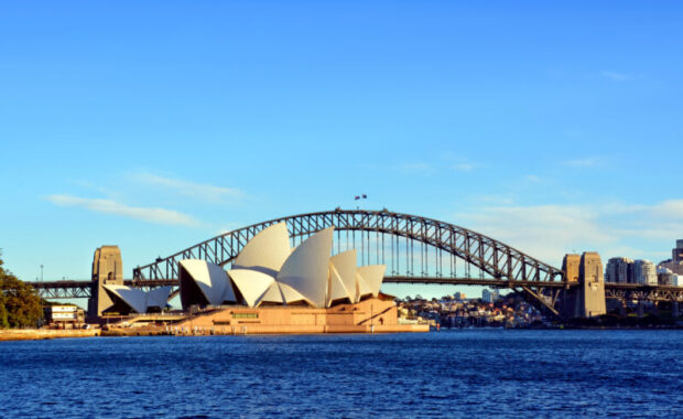 Sydney Opera House with its distinctive sails is set against the backdrop of the Sydney Harbour Bridge. The sky is clear and the water sparkles.