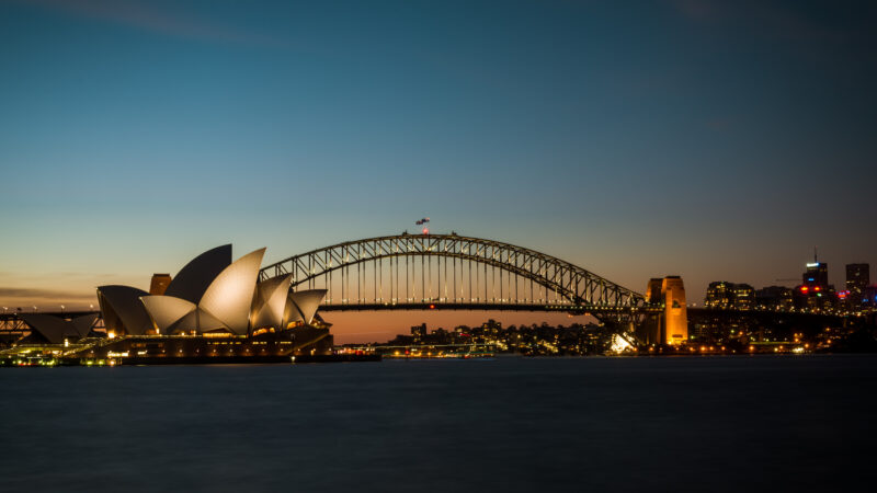 Sunset view of Sydney Harbour, featuring the illuminated Opera House and Harbour Bridge. The city skyline glows softly, creating a serene atmosphere.