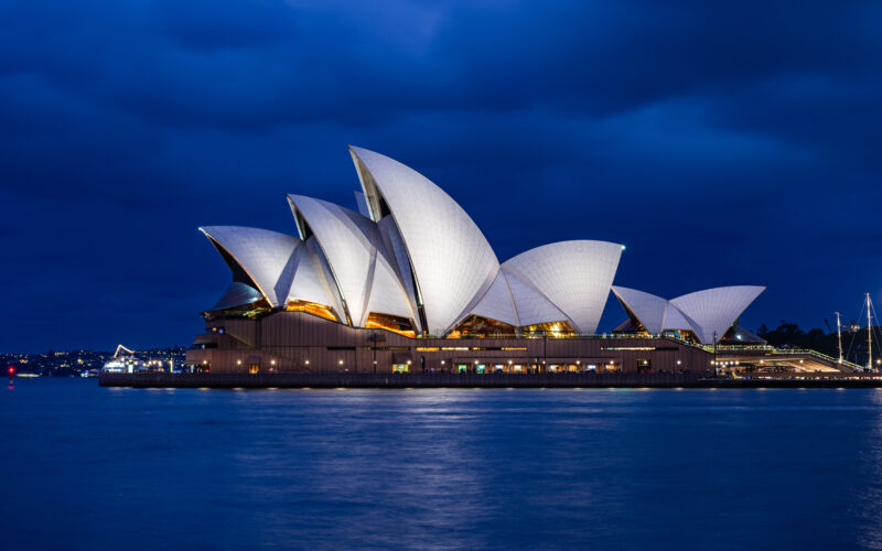 Sydney Opera House illuminated at night, its white sails glowing against a deep blue sky. The calm harbor reflects the lights, creating a serene atmosphere.