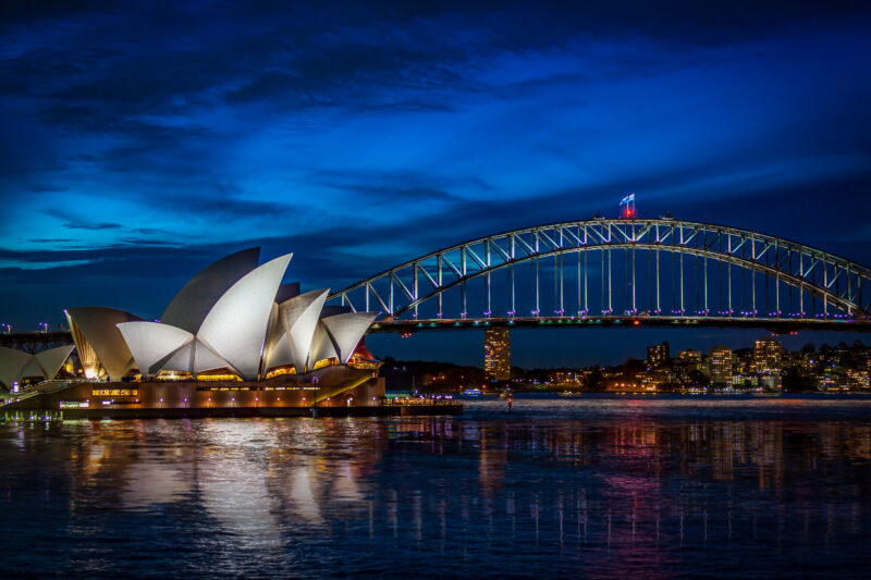 Night view of the Sydney Opera House and Harbour Bridge, illuminated against a deep blue sky. Reflections shimmer on the water, creating a serene scene.