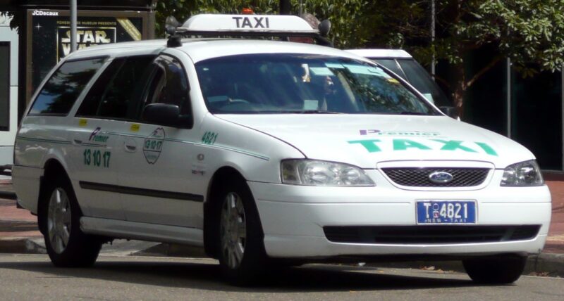 How Far is Sydney Airport from the City? 2 White Sydney station wagon taxi parked on the street. The taxi has a roof sign and displays numbers and a logo on the side. Trees and buildings in the background.