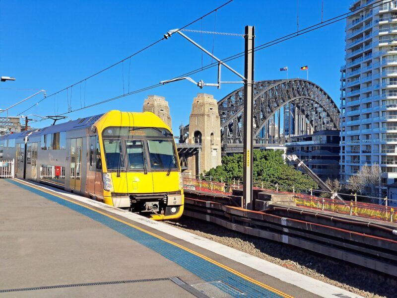 How Far is Sydney Airport from the City? 1 Yellow commuter train is stopped at a sunny station, with Sydney Harbour Bridge prominently in the background under a clear blue sky.