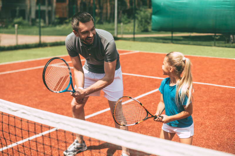 Cheerful father in sports clothing teaching his daughter to play tennis 