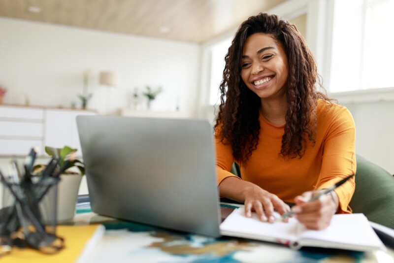 Portrait of smiling woman sitting at desk, using laptop and writing in notebook